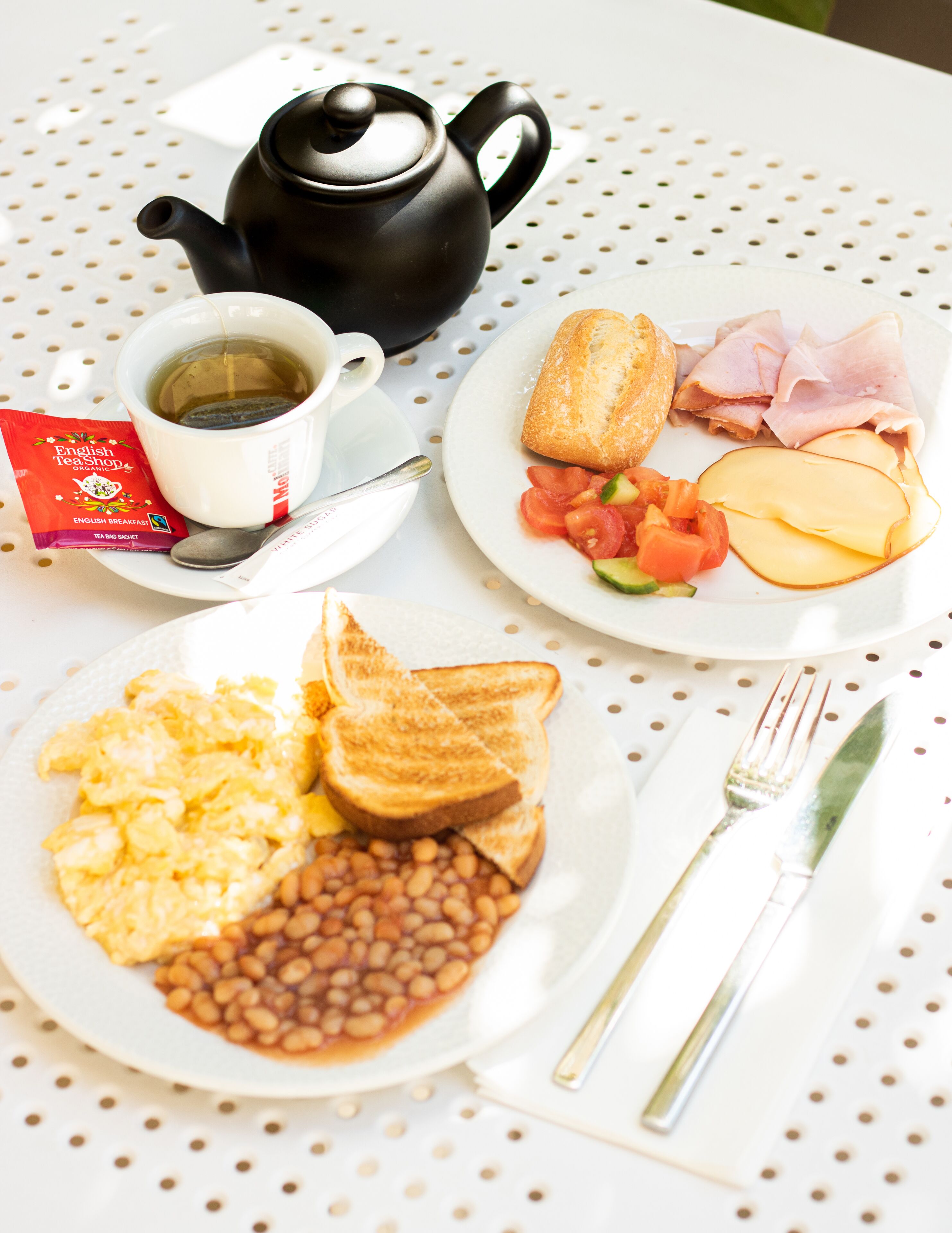 Un desayuno con tetera, taza, tostadas, huevos, frijoles, jamón, queso y tomates en platos.