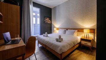 A hotel room with a bed, a desk with a laptop, a chair, a bedside table with a lamp, and a picture on the wall.