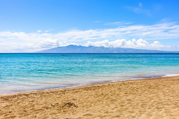 Ein Strand mit klarem türkisblauem Wasser, sandigem Ufer und Bergen in der Ferne unter blauem Himmel.