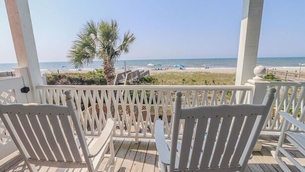 Une terrasse en bois avec deux chaises blanches et une table, donnant sur une plage bordée de palmiers et parsemée de parasols.