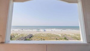 Une vue à travers une fenêtre sur une plage, avec une promenade en bois qui mène au bord de l’eau.