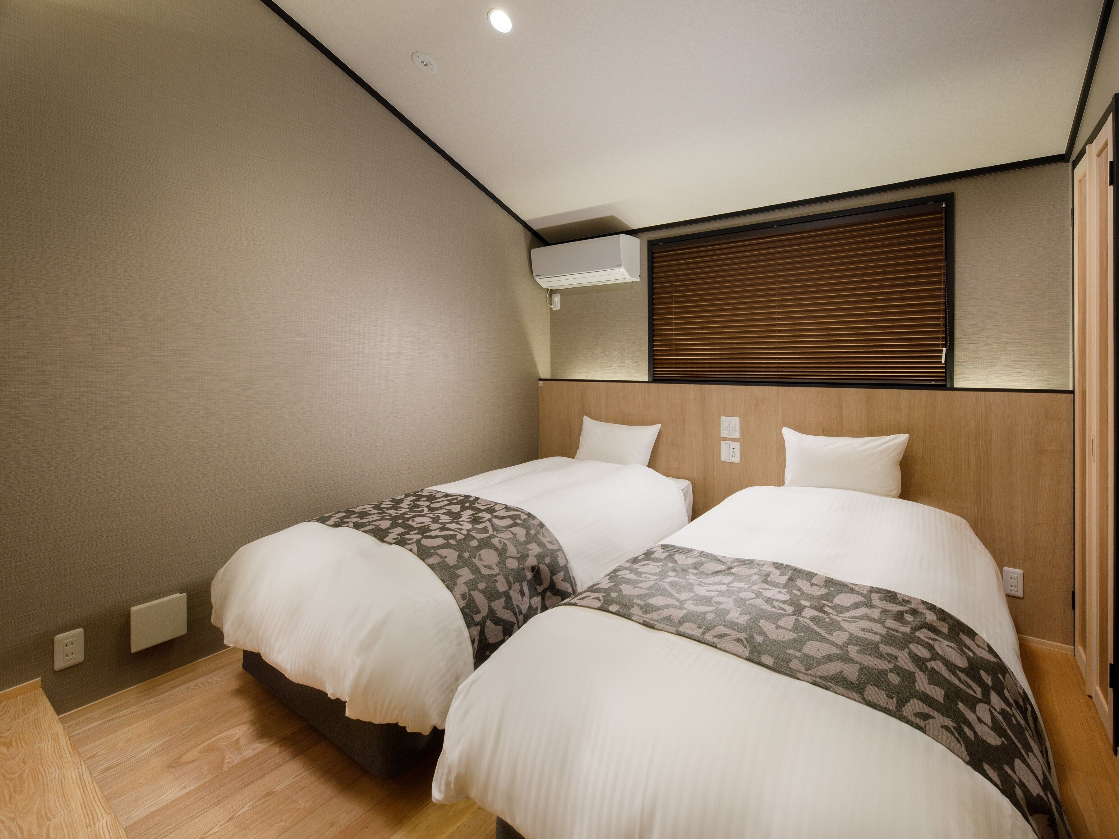 Habitación de hotel con dos camas, cabecera de madera, una lámpara en el techo y una ventana con persianas.