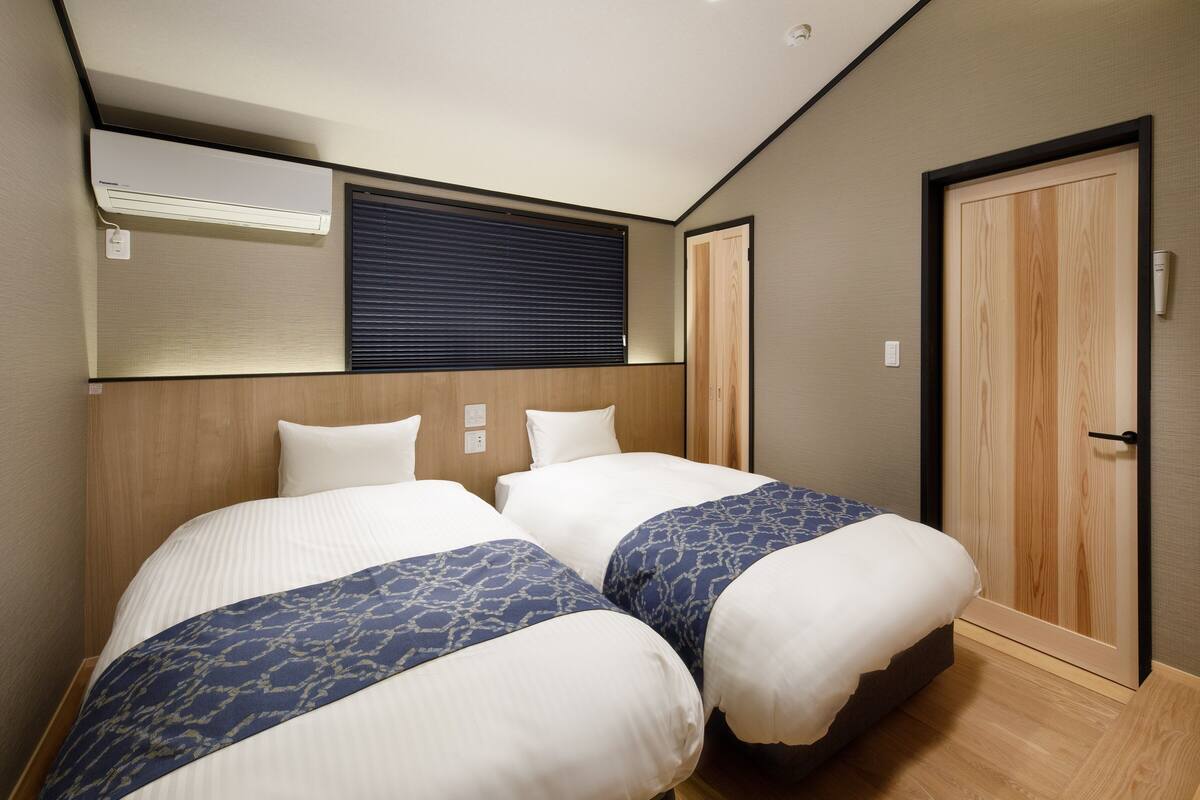 A hotel room with two beds, a wooden wardrobe, a ceiling light, and a window with blinds.