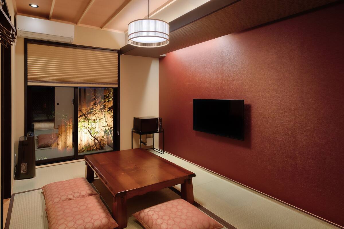 A traditional Japanese room with tatami flooring, a low wooden table, two cushions, a sliding glass door with a view, a mounted TV, and a ceiling light fixture.