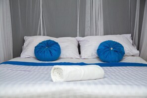 A neatly made bed with white bedding and two blue pillows, a rolled white towel, and sheer curtains in a hotel room.