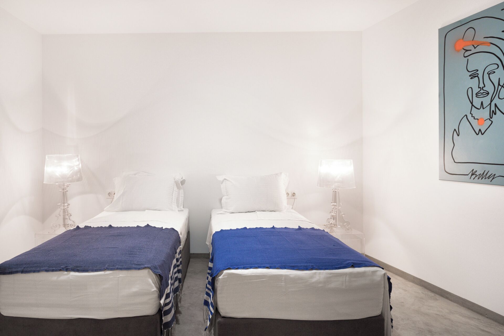 A room with two beds, each with a blue blanket and white pillows, and a wall-mounted lamp.