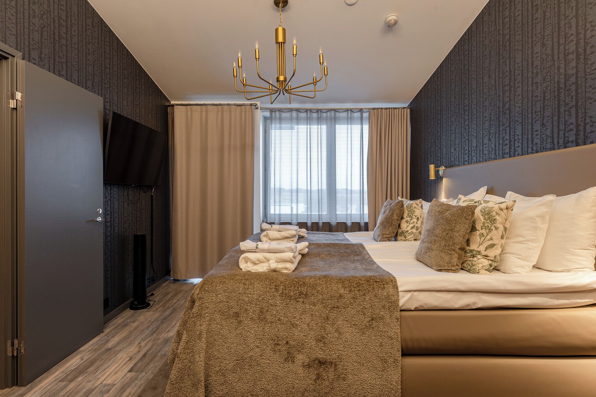 A hotel room with a bed, a flat-screen TV, a chandelier, a large window with curtains, and a wooden floor.