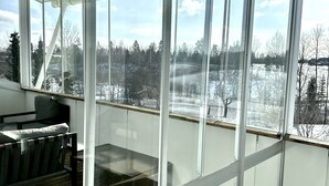 Aurinkoinen huone, jossa on suuret ikkunat, puinen terassi ja näkymä lumiseen maisemaan.