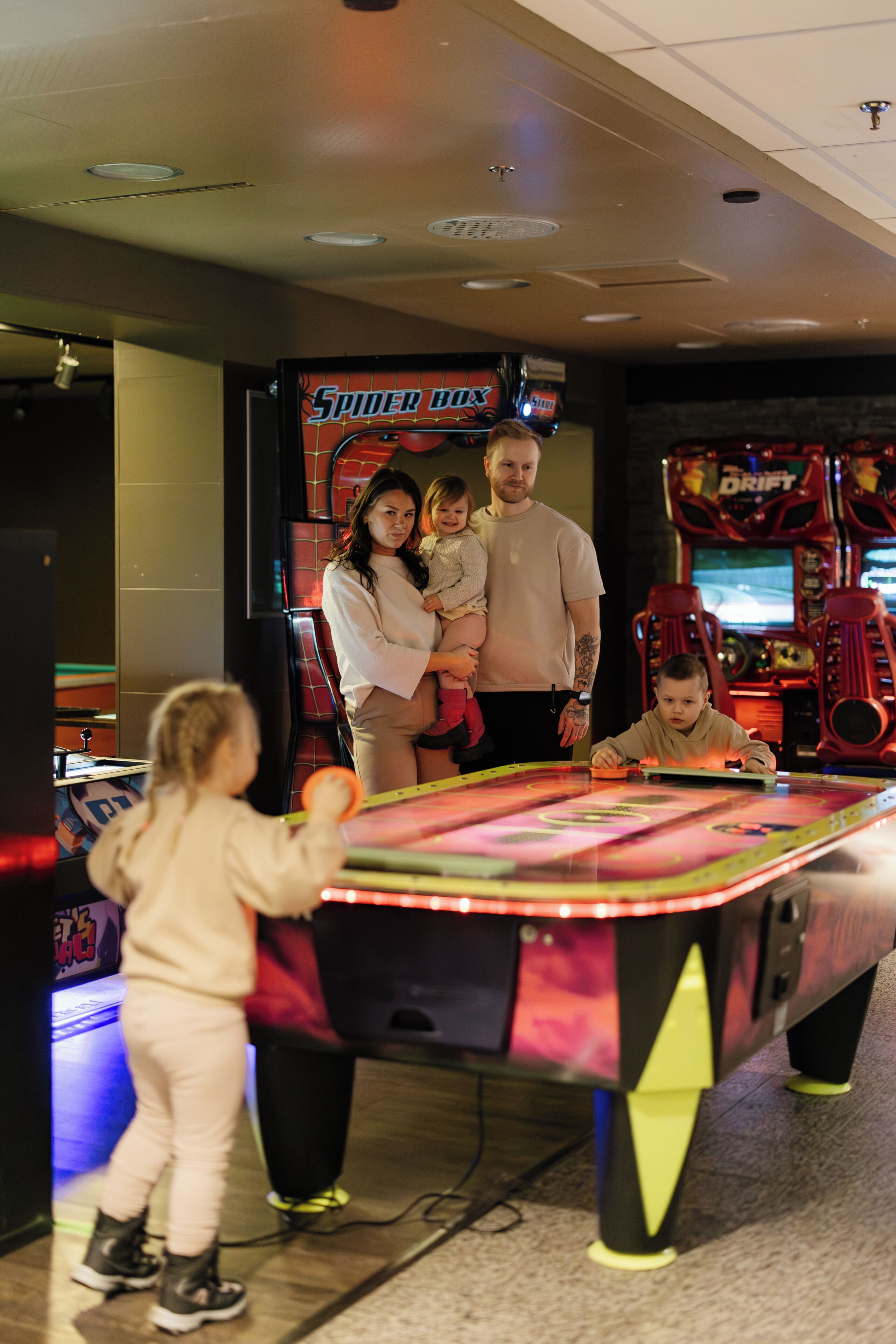 Una familia jugando un juego de mesa en un espacio interior, con máquinas de videojuegos al fondo.