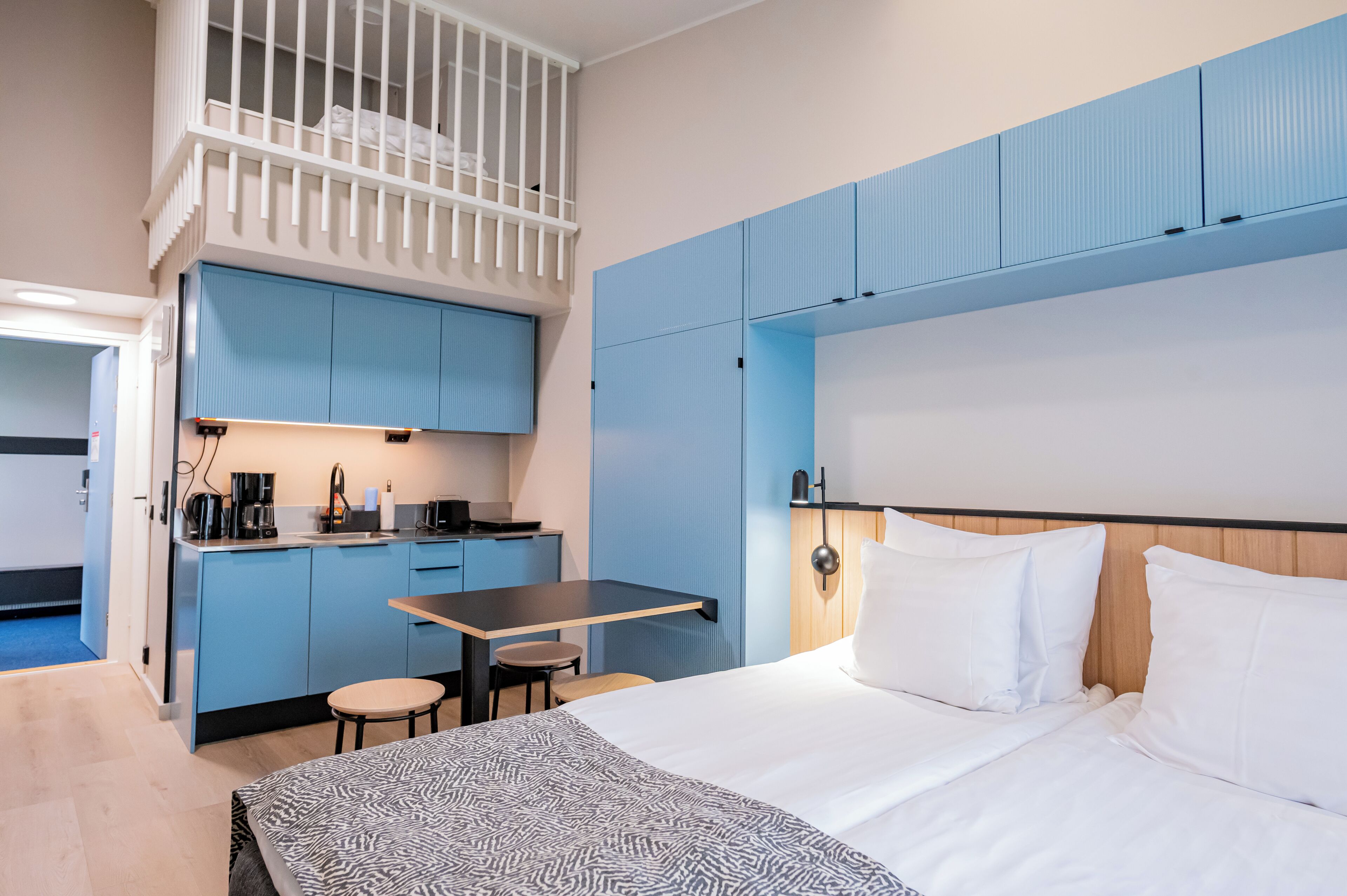 Hotellihuone, jossa on kerrossänky, työpöytä tuolilla, pieni keittotila ja sänky valkoisilla vuodevaatteilla.