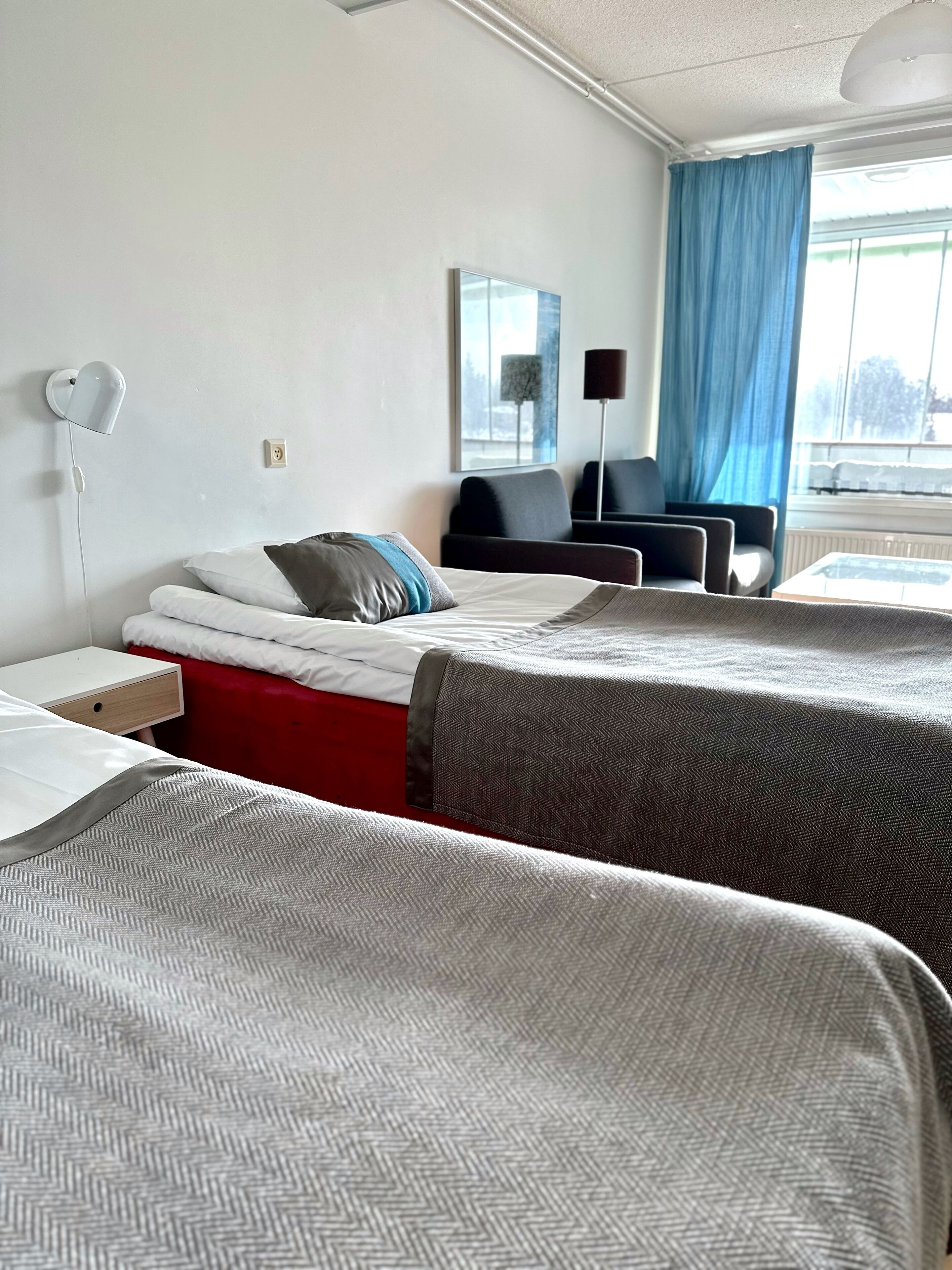 A hotel room with two beds, a desk, a chair, a lamp, and a window with curtains.