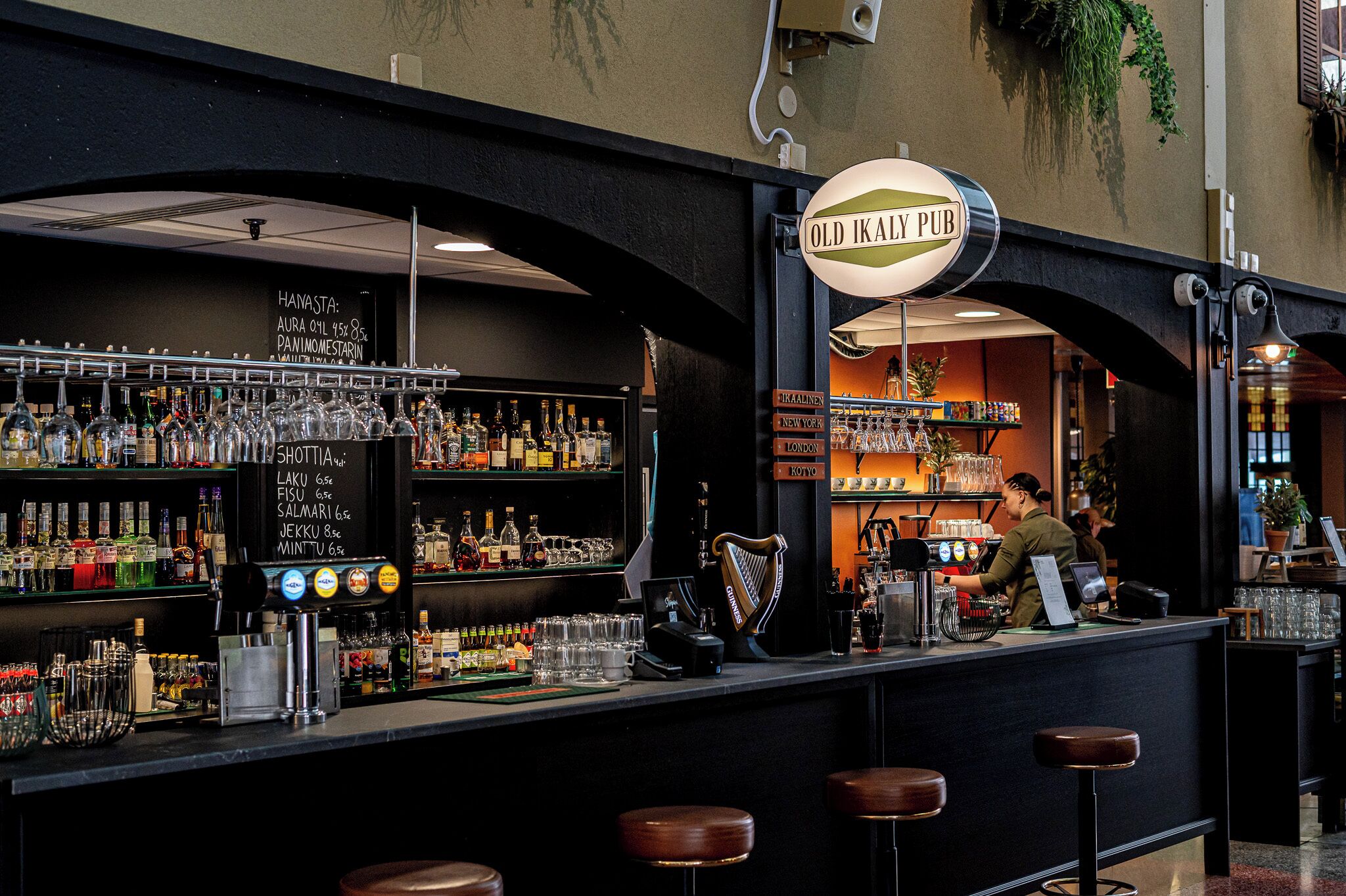 A bar area with a sign reading 'Old Ikaly Pub,' shelves stocked with bottles, a cash register, and stools.
