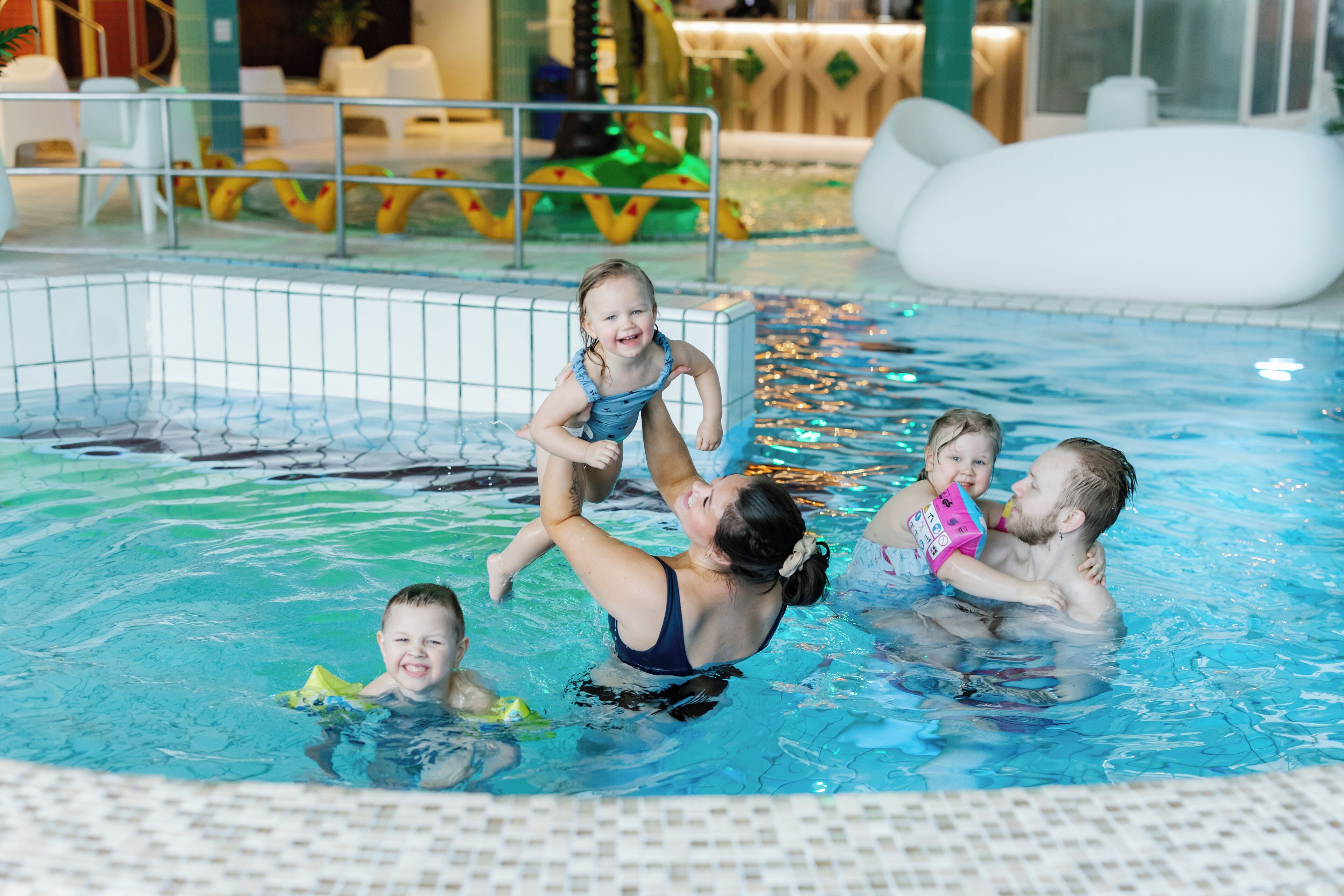 Una piscina con suelo de baldosas, una valla de seguridad y algunas personas nadando.