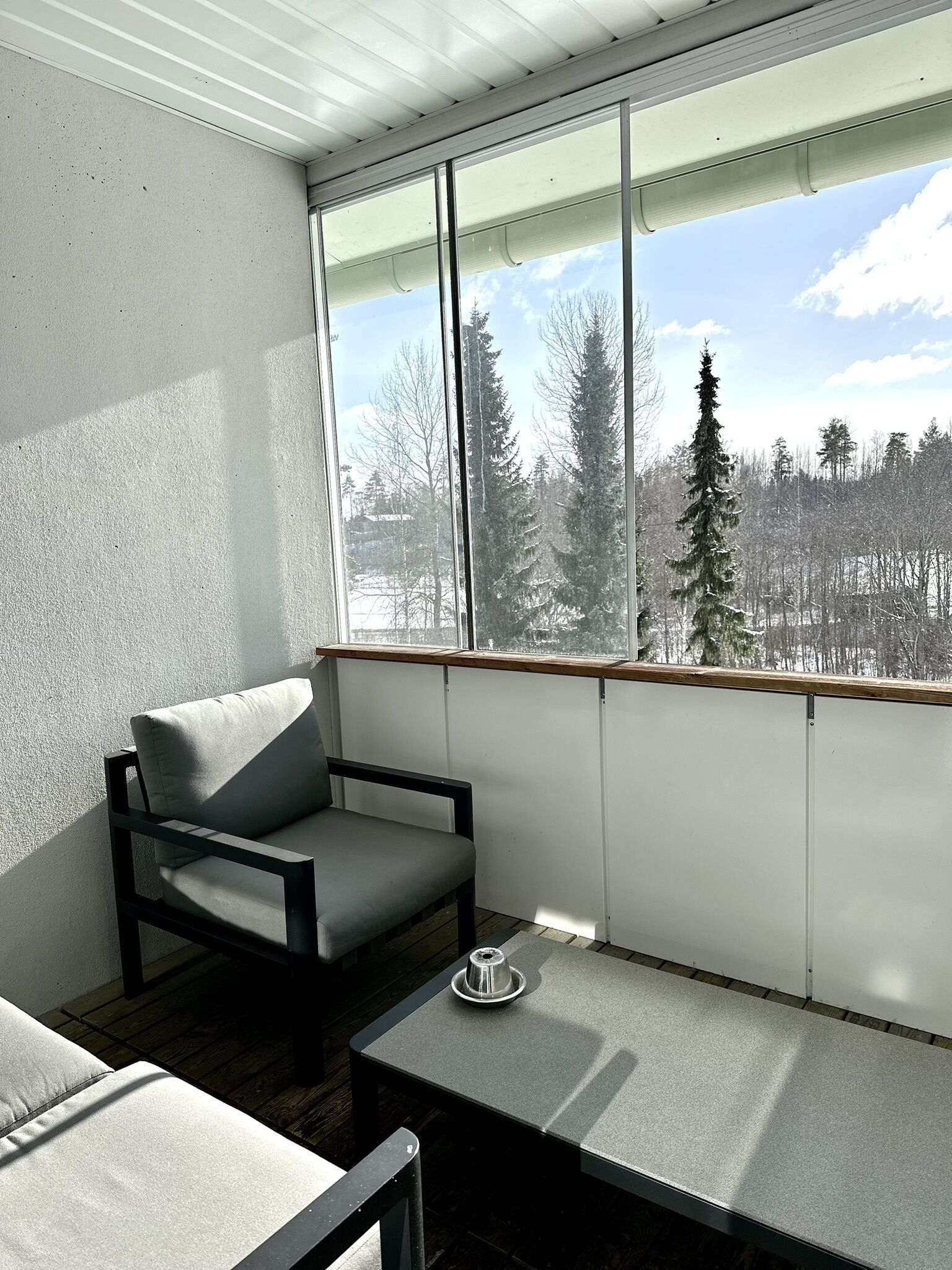 A modern balcony with a grey sofa and a small table, overlooking a snowy forest.