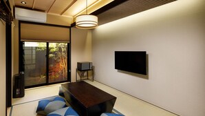 Una habitación con esteras de tatami, una mesa baja, cojines, un televisor y un puertas corredera con vistas a vegetación.