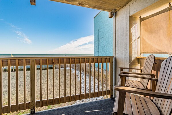 A wooden deck with a view of the beach, outdoor seating, and a building with a blue exterior.