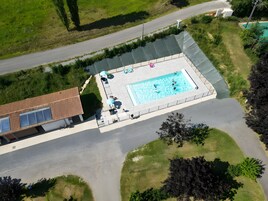 Vue aérienne d’un espace piscine avec une terrasse couverte, un bâtiment doté d’un toit en pente et une route à proximité.