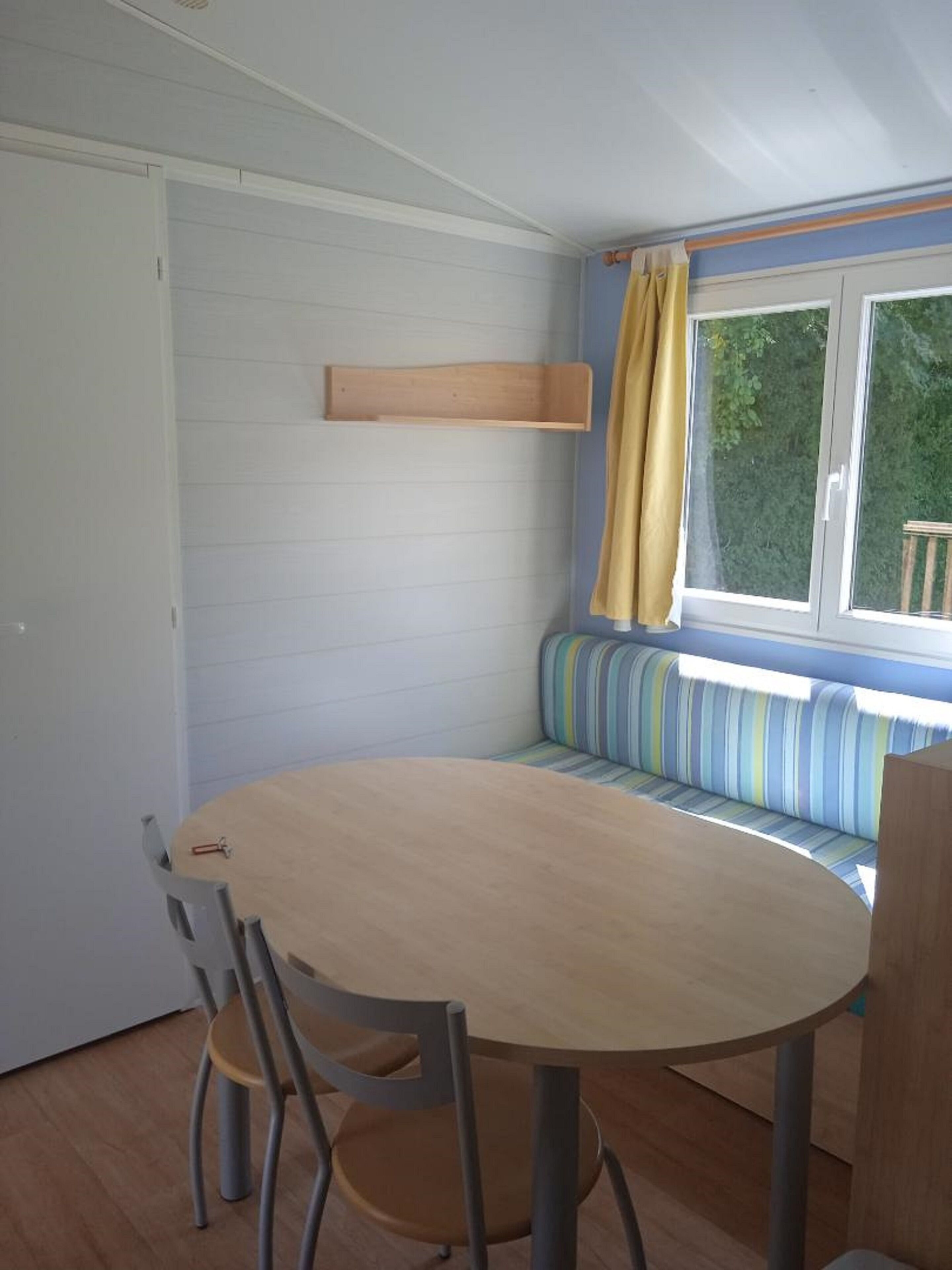 A room with a round table, two chairs, a bench with striped cushions, a wooden shelf, and a window with curtains.