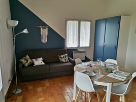 Un coin repas avec une table ronde dressée pour quatre personnes, un canapé de couleur sombre agrémenté de coussins à motifs, une décoration murale en macramé et une fenêtre ornée de rideaux blancs.