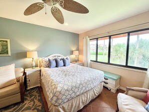 A bedroom with a bed, two bedside tables, a ceiling fan, a window with a view of palm trees, and a small bench. - OV 921 Golf Course View Condo - Paradise Rental in Ocean Village (FORT PIERCE)