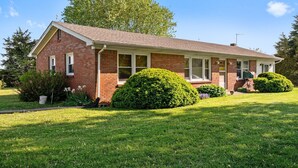 A two-story brick house with a front porch, a small front yard with a well-maintained lawn, and a large tree in the background.