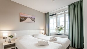 A hotel room with two beds, a nightstand with a lamp, a painting on the wall, and a window with curtains.