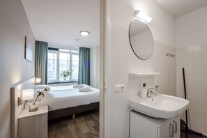 Quarto de hotel moderno com cama, mesa de cabeceira, abajur e uma janela grande com cortinas.