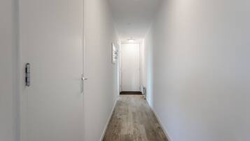 A narrow hallway with white walls and a wooden floor, leading to a door at the end.