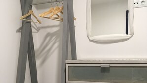 A room with a grey metal shelf unit with hanging clothes, a white mirror, and a grey storage unit with drawers.