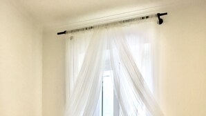 A bedroom with a bed covered in a beige sheet, a window with sheer white curtains, and a white wall.