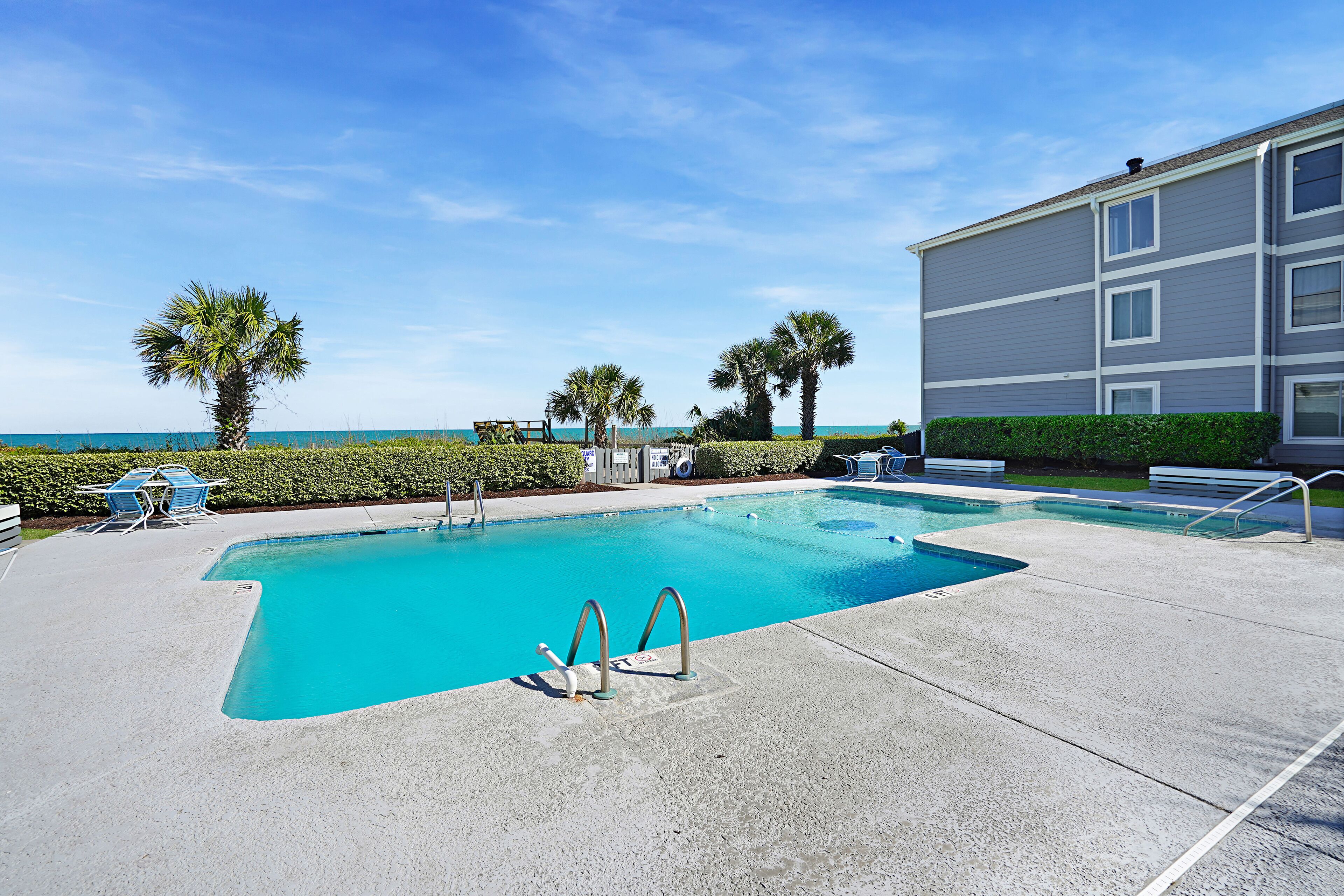 A swimming pool with a clear blue water, surrounded by a concrete deck, palm trees, and a building with a grey facade.