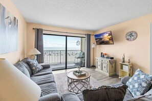 Una sala de estar con sofá, sillón, mesa de centro y vista a la playa a través de un ventanal corredero.