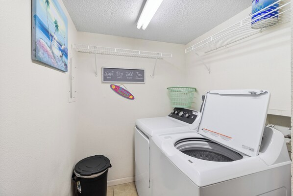 Un cuarto de lavandería con una lavadora de carga frontal, un tendedero, un cubo de basura y un letrero en la pared.