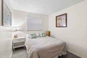 Un dormitorio con cama, mesita de noche, lámpara, ventana con persianas e imagen enmarcada en la pared.