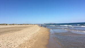En bred sandstrand med milda vågor, en klarblå himmel och avlägsna landformationer.