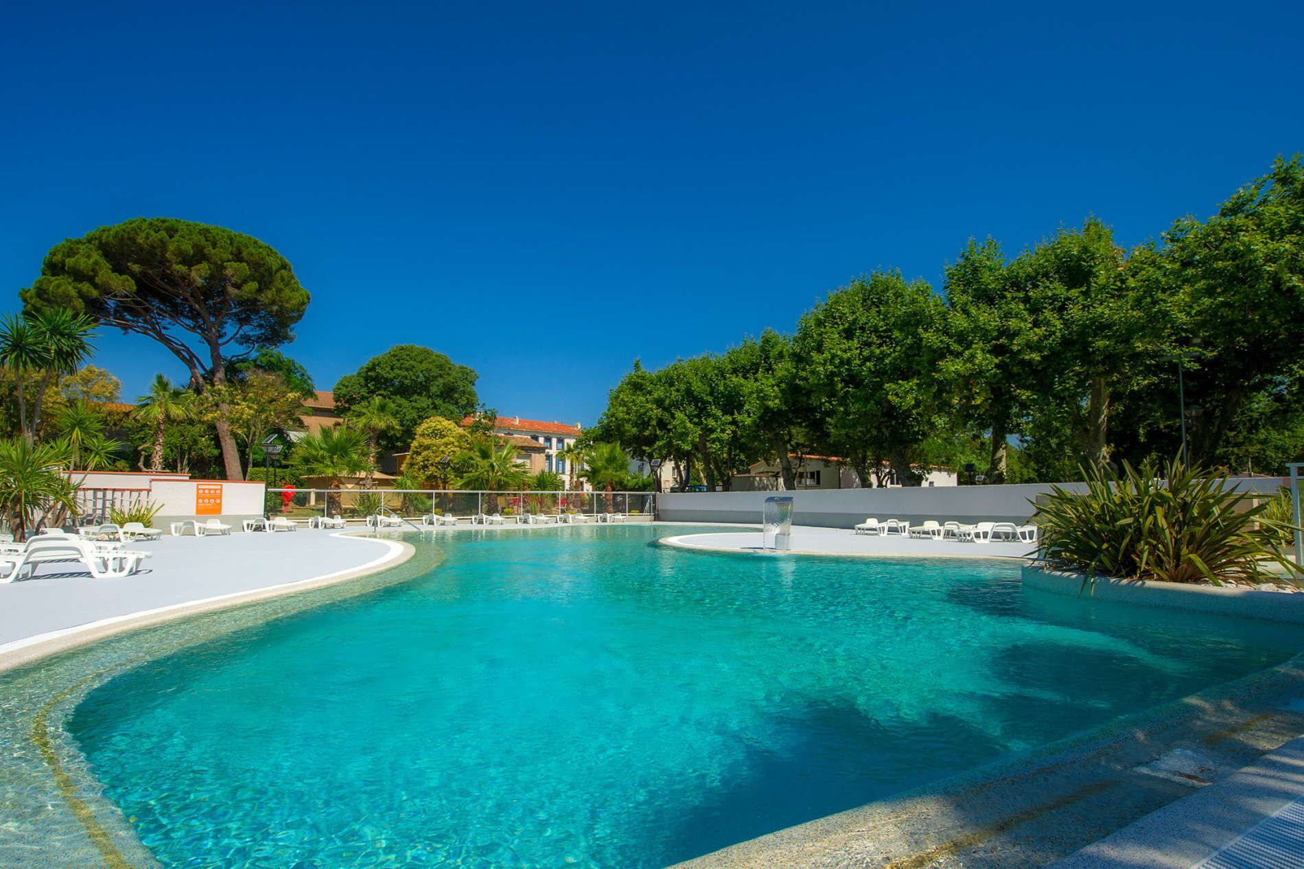 A clear blue pool with lounge chairs and a fountain, surrounded by trees and buildings.
