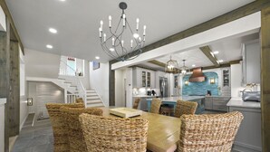 A dining area with a wooden table and wicker chairs, a chandelier, and a kitchen in the background. - Remodeled 5BR Beachfront Home w/ new main floor deck, Private Beach, Communal Pool & Bunks (Santa Rosa Beach)