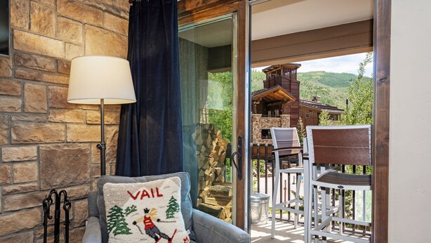 A cozy room with a stone wall, a grey sofa, a white lamp, and a view of a wooden cabin through a glass door. - Heart of Lionshead + Free Parking - Rooftop Hot Tub (Vail)