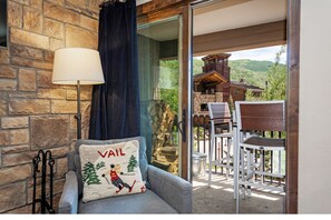 A cozy room with a stone wall, a grey sofa, a white lamp, and a view of a wooden cabin through a glass door.