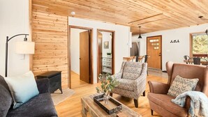 Un salon moderne avec un plafond et des murs en bois, un canapé, des fauteuils, une table basse et un coin repas visible à travers une porte ouverte.