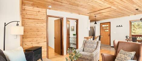 A modern living room with wooden ceiling and walls, a sofa, armchairs, a coffee table, and a dining area visible through an open door.