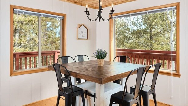 Un espace repas comprenant une table et des chaises en bois, un lustre et de grandes fenêtres donnant sur la forêt.