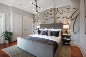 A bedroom with a large bed, a nightstand, a lamp, a potted plant, and a wall with abstract art.