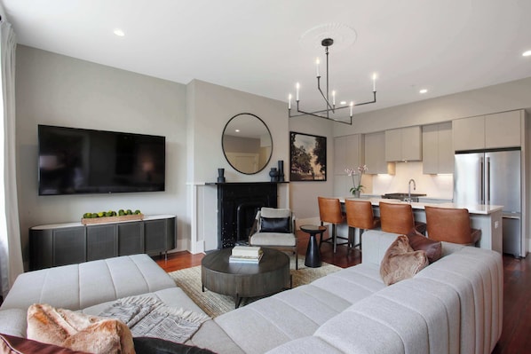 A modern living room with a sofa, coffee table, and TV. There is a fireplace, dining area, and kitchen with a refrigerator.