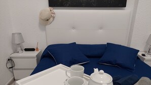 Une chambre d’hôtel bien rangée, avec un lit, une table de chevet dotée d’une lampe, un plateau avec une théière et des tasses, et un chapeau accroché au mur.