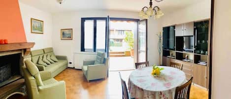 A living room with a sofa, a dining table with chairs, a kitchen area with cabinets, and a large window.