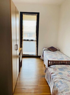 Een smalle kamer met houten vloer, een bed met beddengoed met patroon, en een deur naar een balkon.