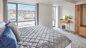 Une chambre avec une grande fenêtre, un lit recouvert d’une courtepointe matelassée, une table de chevet avec une lampe, et une vue sur un port de plaisance.