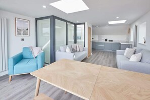 A modern living room with a large wooden table, two sofas, and a bright kitchen in the background.