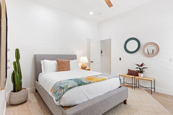 Un dormitorio moderno con cama, mesita de noche, lámpara, planta de cactus y espejo en la pared.